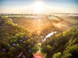 Aerial view of small cabins surrounded by trees near a lake with the sun rising over fields at Deerpark Classic Golden Oak Treehouse in Deerpark Forest