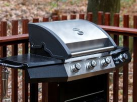 A gas grill with four control knobs and cooking utensils hanging on the side on a wooden deck at Strathyre Golden Oak Hideaway in Strathyre Ben Ledi