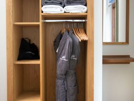 A wooden wardrobe with towels on the top shelf, bathrobes hanging on wooden hangers, a hairdryer in a black bag in a cubby hole next to a mirror with a wooden shelf