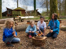 Four children roasting marshmallows on sticks over a fire pit in a wooded outdoor area with log seating and wooden buildings in the background at Forest of Dean Meadow - Golden Oak in Forest Of Dean