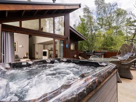 An outdoor deck with a hot tub wicker chairs a glass table and barbecue grill next to a cabin with large windows and open doors at Glentress Forest White Willow Premium Glentress Forest Peebles
