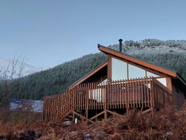 A wooden cabin with a fenced porch set against a backdrop of forested hills with some snow at Strathyre Golden Oak Hideaway in Strathyre Ben Ledi
