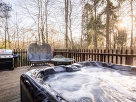 An outdoor wooden deck with a hot tub a grill a wicker swing chair and a table surrounded by trees at Forest of Dean Golden Oak Hideaway in Forest Of Dean