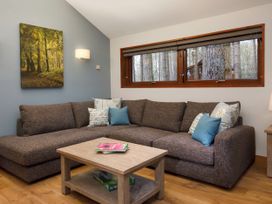 A living room with a brown sectional sofa with cushions a wooden coffee table with books and board games a window showing trees and a wall painting at Strathyre New Style Silver Birch in Strathyre Ben Ledi