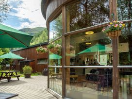 An outdoor seating area with picnic tables and green umbrellas next to a building with large glass windows and hanging flower baskets at Strathyre New Style Golden Oak in Strathyre, Ben Ledi