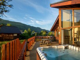 A wooden deck with a hot tub picnic table and barbecue grill outside a cabin with mountains in the background at Ardgartan Argyll Golden Oak Lochside in Ardgartan Loch Long