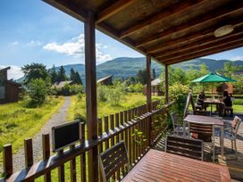 A wooden patio with tables and chairs and a green umbrella with people sitting and mountains in the background at Ardgartan Argyll Golden Oak Lochside Ardgartan Loch Long
