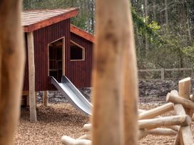 A wooden playhouse with a metal slide in an outdoor playground with wood chips on the ground and a wooded area in the background at Delamere Forest Golden Oak in Delamere Forest