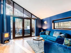 A living room with blue walls two blue sofas a round wooden coffee table a rug a wood-burning stove and large glass doors opening to a deck at Garwnant Golden Oak Hideaway in Garwnant Bannau Brycheiniog Brecon Beacons
