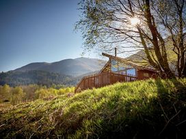 A wooden cabin on a grassy hill with trees and mountains in the background at Strathyre Golden Oak Hideaway Lochside Strathyre Ben Ledi