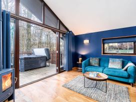 A living room with a teal sofa blue walls a wood stove and large glass doors leading to a deck with a hot tub at Glentress Forest Golden Oak Hideaway in Glentress Forest Peebles