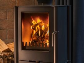 A wood burning stove with fire inside and a basket of firewood nearby at Forest of Dean Golden Oak Hideaway in Forest Of Dean