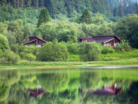 Two wooden houses surrounded by trees near a lake with reflections in the water at Strathyre Classic Golden Oak Lochside Strathyre Ben Ledi