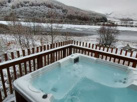 An outdoor hot tub on a wooden deck overlooking a snowy landscape with trees and a partially frozen lake at Strathyre Classic Golden Oak Lochside in Strathyre Ben Ledi
