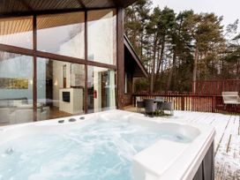 An outdoor deck with a hot tub table and chairs and a barbecue grill next to a house with large glass windows and a forest in background at Cropton Silver Birch Compact Cropton Forest