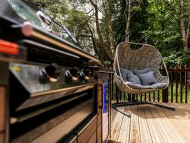 An outdoor area with a barbecue grill and a hanging wicker chair with cushions on a wooden deck surrounded by trees at Keldy White Willow Premium in Keldy Forest