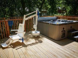 An outdoor wooden deck with a hot tub and a mounted chair lift next to a wooden railing surrounded by trees at Beddgelert Silver Birch - freshly inspired design wheelchair accessible cabin in Beddgelert Eryri Snowdonia