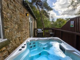 A hot tub on a wooden deck next to a stone wall with a barbecue grill and table with chairs at Keldy Golden Oak Cottage in Keldy Forest