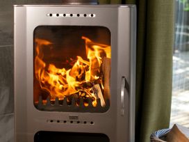 A metal wood-burning stove with fire inside next to a window with green curtains at Cropton Golden Oak in Cropton Forest