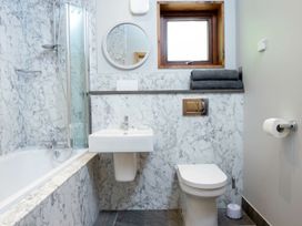 A bathroom with marble walls featuring a bathtub shower combination a wall-mounted sink a toilet with a toilet brush and holder a round mirror a window and folded towels on a shelf at Garwnant White Willow Premium Valley View in Garwnant Bannau Brycheiniog Brecon Beacons