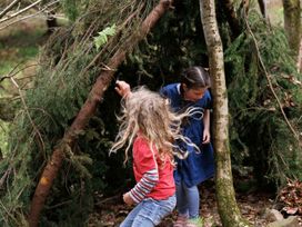 Two children playing near a structure made of tree branches and leaves in a forest