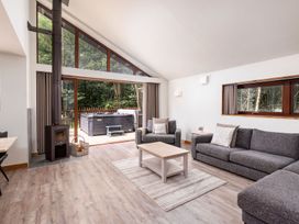 A living room with grey sofas a wooden coffee table a wood burning stove and large windows overlooking a deck with a hot tub at Glentress Forest White Willow Premium in Glentress Forest Peebles