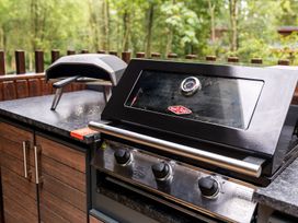 An outdoor kitchen counter with a grill and a portable pizza oven with trees and wooden fence in the background at Glentress Forest White Willow Premium Glentress Forest Peebles