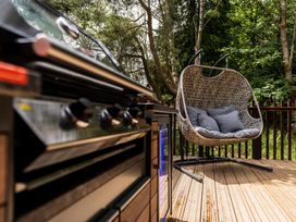 An outdoor deck with a barbecue grill and a wicker hanging chair with cushions in a wooded area at Glentress Forest White Willow Premium in Glentress Forest Peebles