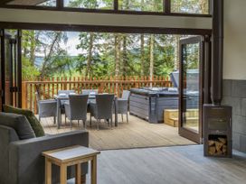 A living room with a wood stove and a small wooden table next to a grey sofa opening onto a deck with a dining table and chairs and a hot tub surrounded by trees at Glentress Forest White Willow Premium Valley View Glentress Forest Peebles