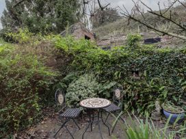A small outdoor patio with a metal table and two chairs surrounded by dense greenery and a brick wall at Anvil Cottage in Gatcombe near Blakeney