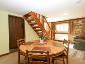 A dining area with a round wooden table and chairs near a staircase and window with curtains at Anvil Cottage in Gatcombe near Blakeney