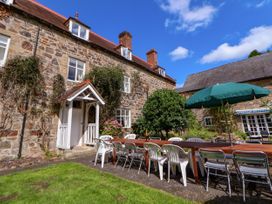 An outdoor patio area with a long wooden table surrounded by white and metal chairs and a green umbrella by a stone house at New Hall in Chirk