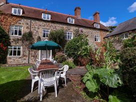 An outdoor patio with a long wooden table and white plastic chairs under a green umbrella in front of a stone house at New Hall in Chirk