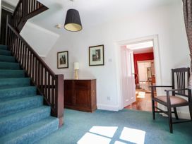 An entryway with a staircase carpeted in blue with dark wood railing a wooden sideboard with a lamp and framed pictures on the wall a wooden chair with cushions and a doorway leading to another room with red walls at New Hall in Chirk