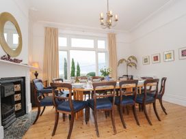 A dining room with wooden floor a large wooden table set with plates and glasses wooden chairs with blue upholstery a fireplace with a round mirror and a lamp near large windows at Duncombe in Teignmouth