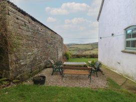 An outdoor seating area with a table and benches between a stone wall and a white building overlooking fields at Priddbwll in Llansannan