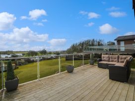 A wooden deck with a brown cushioned outdoor sofa and potted plants overlooking a grassy area and buildings at 1 Fishermans Hill in Glenluce