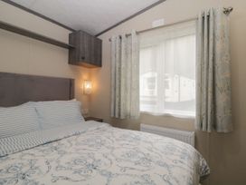 A bedroom with a bed with patterned bedding striped pillows a wall light window with floral curtains and a cabinet at Caravan 7 in Plymouth