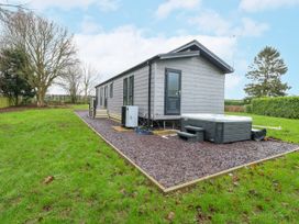 A small gray house with multiple windows and a door next to a hot tub on gravel surrounded by grass and trees at Wurzburc in Swaffham