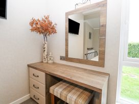 A wooden dressing table with drawers and a plaid cushioned stool a vase with orange leaves and a decorative cage on the table a mirror on the wall reflecting a television a window showing greenery outside at Wurzburc in Swaffham