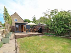A garden with a wooden patio table and chairs under an umbrella a barbecue and a small wooden outbuilding at Bramley Cottage near Cirencester
