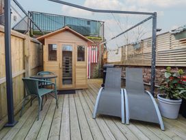 An outdoor deck area with a small wooden shed a round glass table with two chairs two lounge chairs a barbecue grill and potted plants at Circus Circus in Holywell