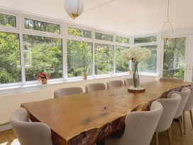 A dining room with a large wooden table surrounded by chairs and windows showing greenery at Serenity House The Retreat in Llangammarch Wells near Llanwrtyd Wells