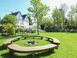 A circular wooden bench seating area around a fire pit in a grassy garden with trees and a house in the background at Serenity House The Retreat in Llangammarch Wells near Llanwrtyd Wells
