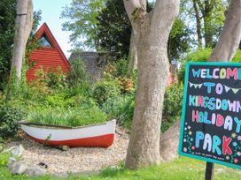An outdoor scene with a small red and white boat used as a planter surrounded by greenery and a colorful welcome sign for Kingsdown Holiday Park