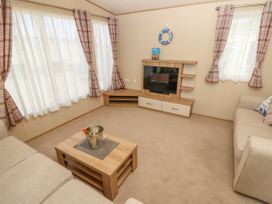 A living room with two beige sofas a wooden coffee table with a wine bottle and ice bucket a corner cabinet with an electric fireplace and three plaid curtain windows at Coastal Cottage in East Wittering
