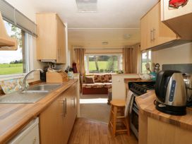 A kitchen with wooden cabinets, a sink, kettle, stove, and a stool leading to a sitting area with a sofa and window showing a green field at Hollie in Raglan