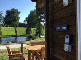 Wooden chairs and table on a deck near a pond with green trees and grass at Hollie Lodge Raglan