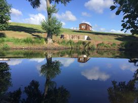 A grassy hillside with a wooden cabin a picnic table and barbecue near a pond reflecting the sky at Poppy in Raglan
