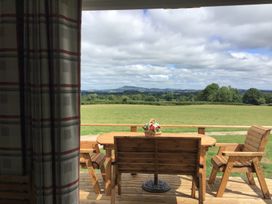 A wooden outdoor table with chairs and a flower basket on a deck overlooking a green field and distant hills at Poppy in Raglan